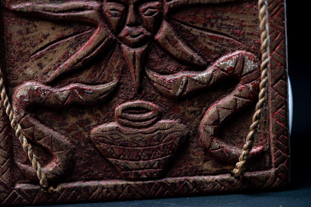 Ancient shaman box from the Paiwan tribe, stained red, with a sliding lid carved with snakes face. One side is carved with two tattooed hands, while the other features a sun, a pot, and two snakes — depicting the myth of the Paiwan people's origin from the union of a pot and a snake. The sides are inlaid with circular shell ornaments.