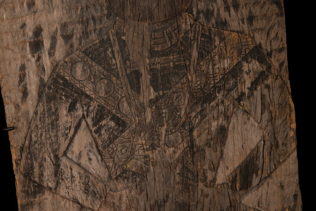 Old ancestor panel from the Paiwan tribe, carved with a standing warrior, one hand resting on his rifle and the other on the pommel of his sword. He is dressed in ceremonial attire and wears a hat adorned with eagle feathers, a symbol of valor.