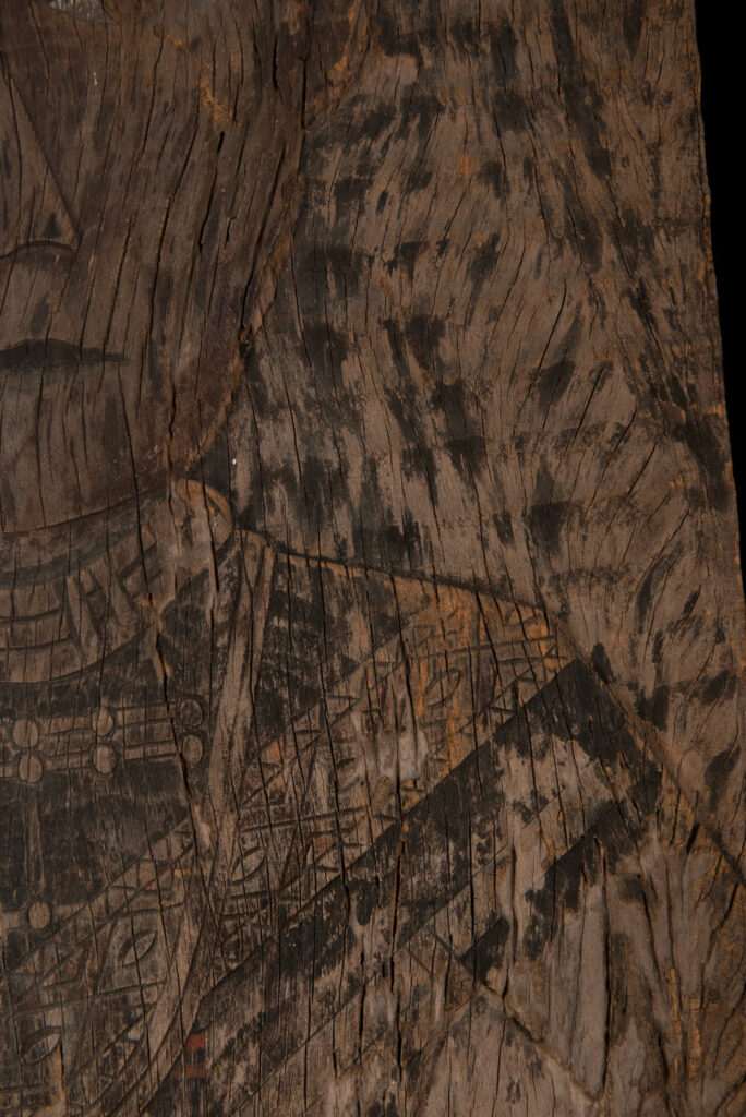 Old ancestor panel from the Paiwan tribe, carved with a standing warrior, one hand resting on his rifle and the other on the pommel of his sword. He is dressed in ceremonial attire and wears a hat adorned with eagle feathers, a symbol of valor.