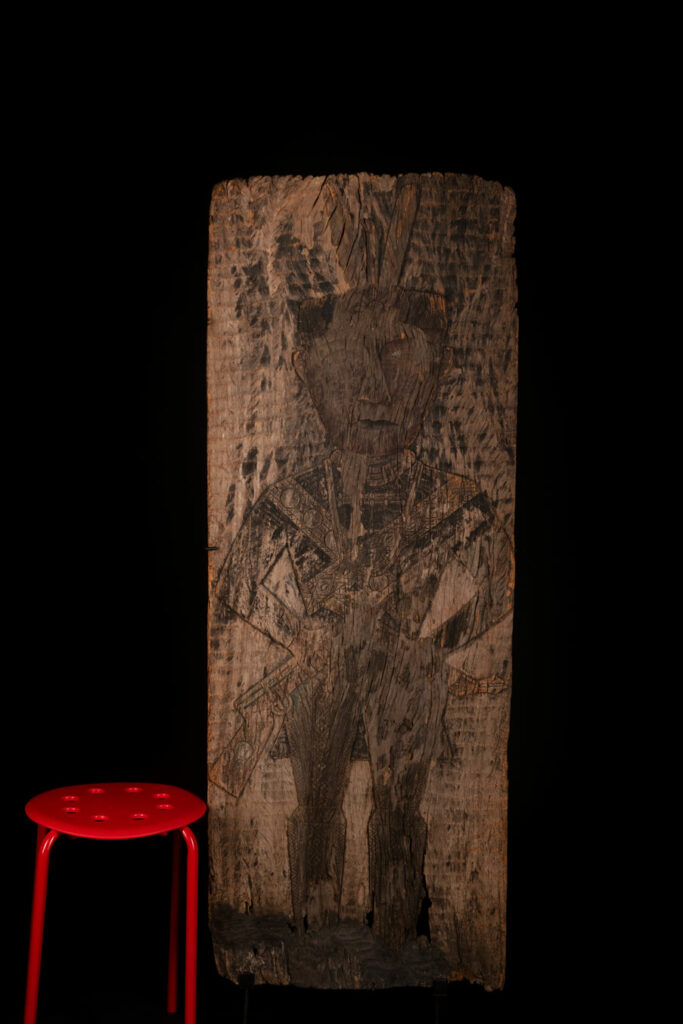 Old ancestor panel from the Paiwan tribe, carved with a standing warrior, one hand resting on his rifle and the other on the pommel of his sword. He is dressed in ceremonial attire and wears a hat adorned with eagle feathers, a symbol of valor.