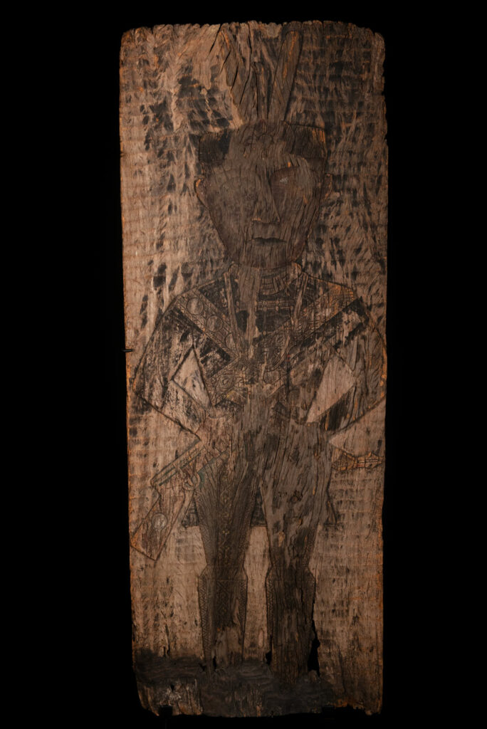 Old ancestor panel from the Paiwan tribe, carved with a standing warrior, one hand resting on his rifle and the other on the pommel of his sword. He is dressed in ceremonial attire and wears a hat adorned with eagle feathers, a symbol of valor.