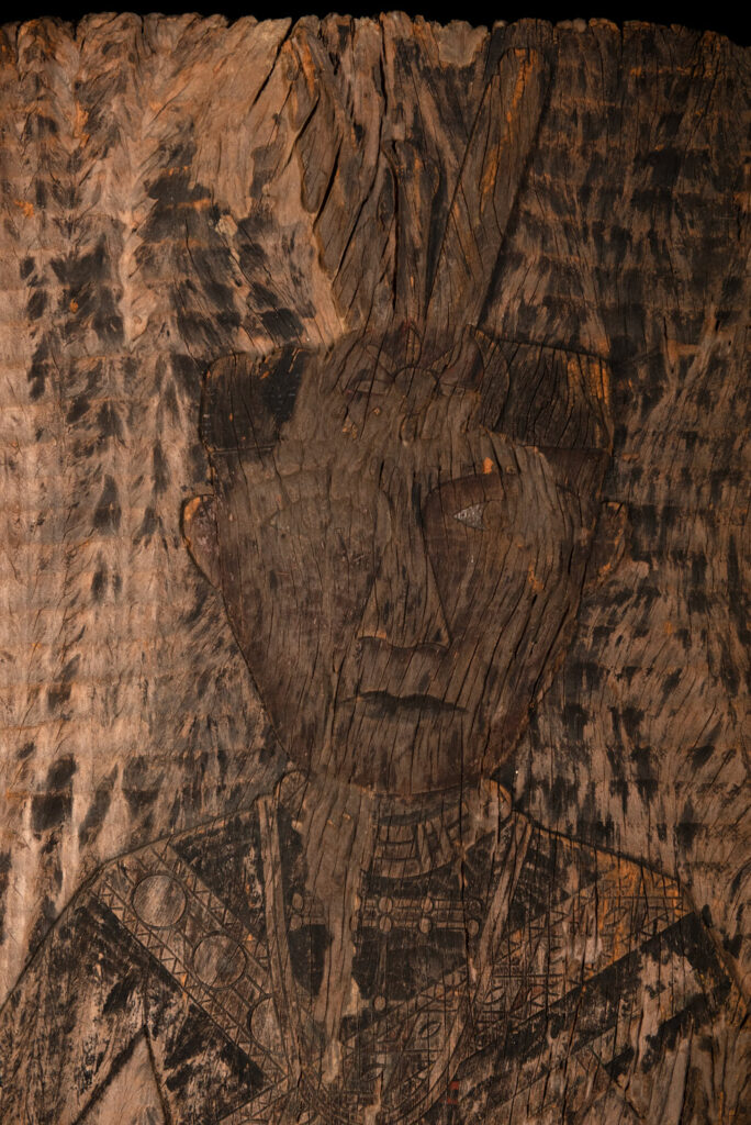 Old ancestor panel from the Paiwan tribe, carved with a standing warrior, one hand resting on his rifle and the other on the pommel of his sword. He is dressed in ceremonial attire and wears a hat adorned with eagle feathers, a symbol of valor.