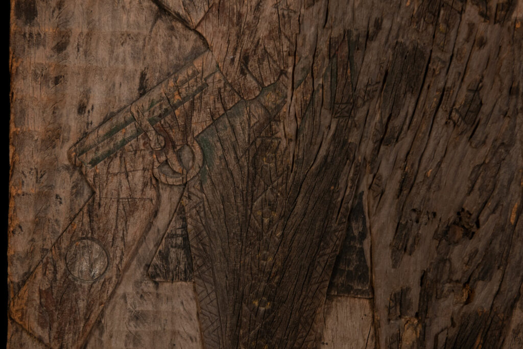 Old ancestor panel from the Paiwan tribe, carved with a standing warrior, one hand resting on his rifle and the other on the pommel of his sword. He is dressed in ceremonial attire and wears a hat adorned with eagle feathers, a symbol of valor.