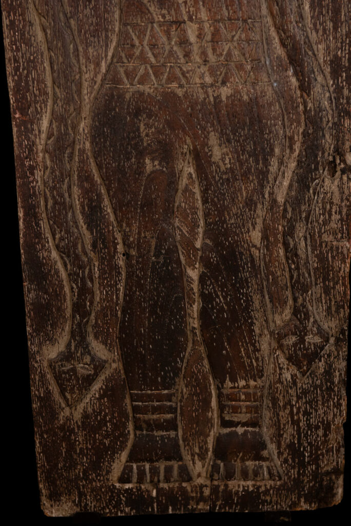 Ancestor panel from the Paiwan tribe, carved with a figure wearing a hat adorned with eagle feathers, a symbol of valor. His face is encircled by two snakes merging into one, and flanked by two additional sacred snakes on each side.