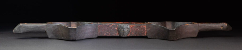 This linked cup from the Paiwan tribe features a diamond-shaped mouth and a central handle adorned with a carved human head. It is decorated with traditional black and red pigments.