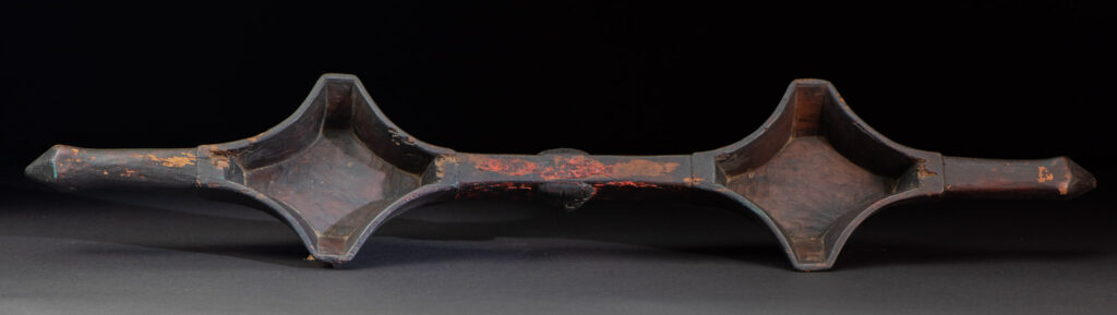 This linked cup from the Paiwan tribe features a diamond-shaped mouth and a central handle adorned with a carved human head. It is decorated with traditional black and red pigments.