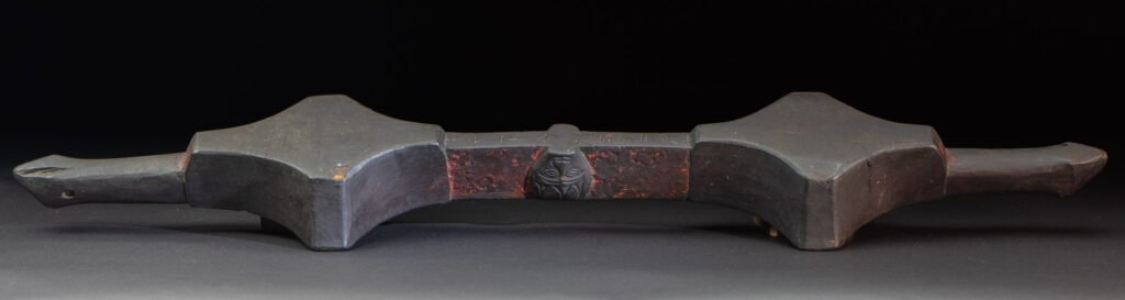 This linked cup from the Paiwan tribe features a diamond-shaped mouth and a central handle adorned with a carved human head. It is decorated with traditional black and red pigments.