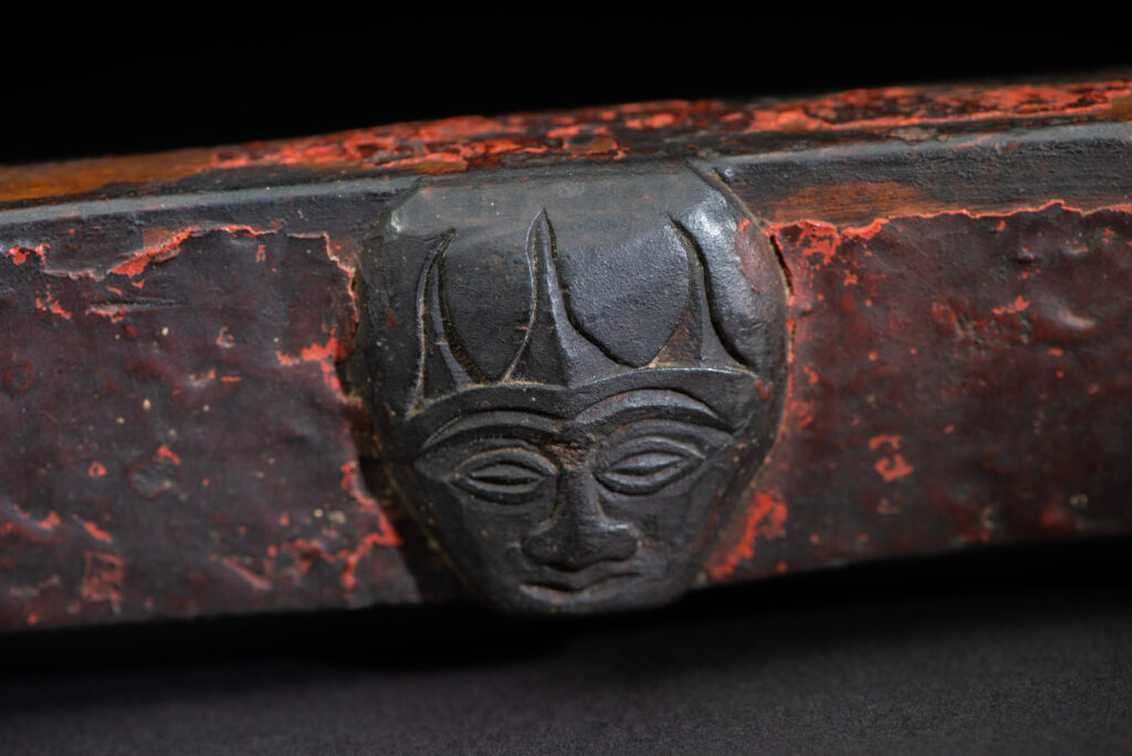 This linked cup from the Paiwan tribe features a diamond-shaped mouth and a central handle adorned with a carved human head. It is decorated with traditional black and red pigments.