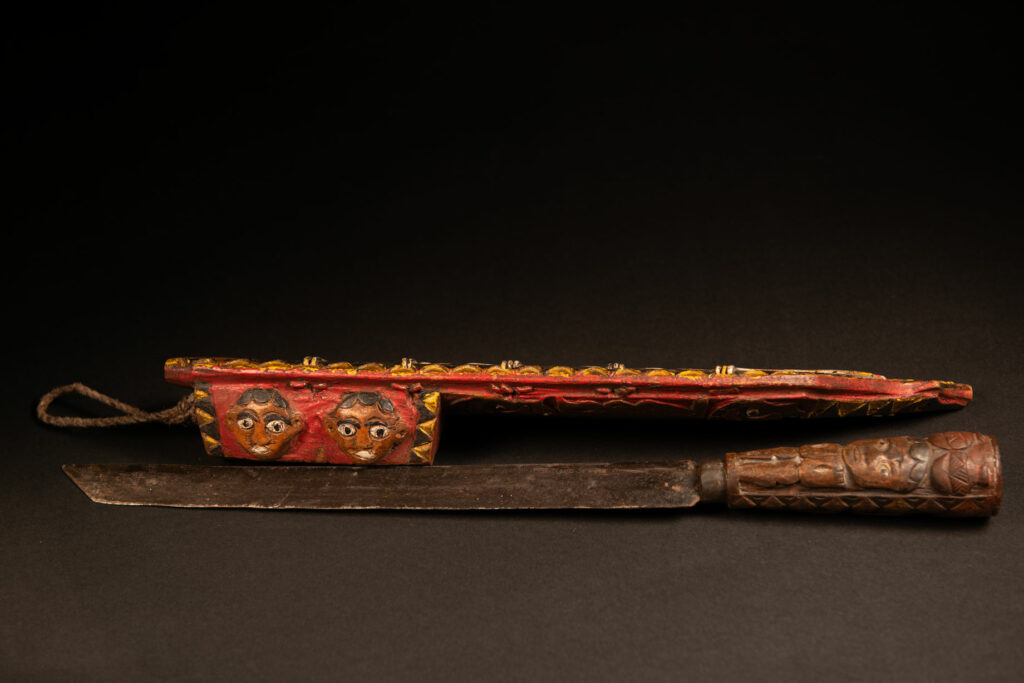 Hunting sword from the Paiwan tribe, carved with a snake and a decorative frieze on the front, as well as a stylized standing figure, a human face, and a snake. The side of the scabbard is carved with two faces. The entire piece is painted in red, brown, white, black, and yellow. The paint was likely applied at a later time.