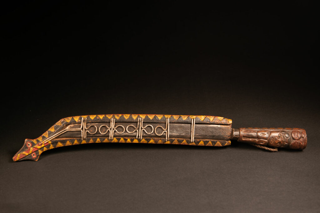 Hunting sword from the Paiwan tribe, carved with a snake and a decorative frieze on the front, as well as a stylized standing figure, a human face, and a snake. The side of the scabbard is carved with two faces. The entire piece is painted in red, brown, white, black, and yellow. The paint was likely applied at a later time.