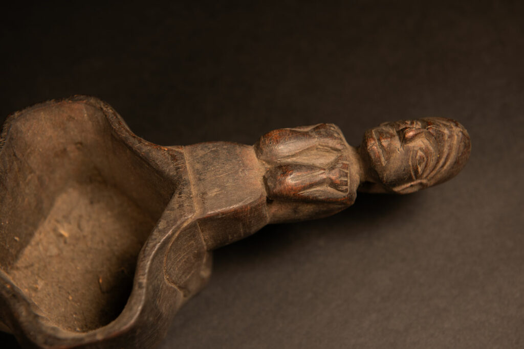 This drinking cup from paiwan features two beautifully carved handles, each depicting an ancestor holding the drinking cup between his thighs. The center of the double cup is carved with four human faces and a decorative frieze. The entire piece is made of natural wood.