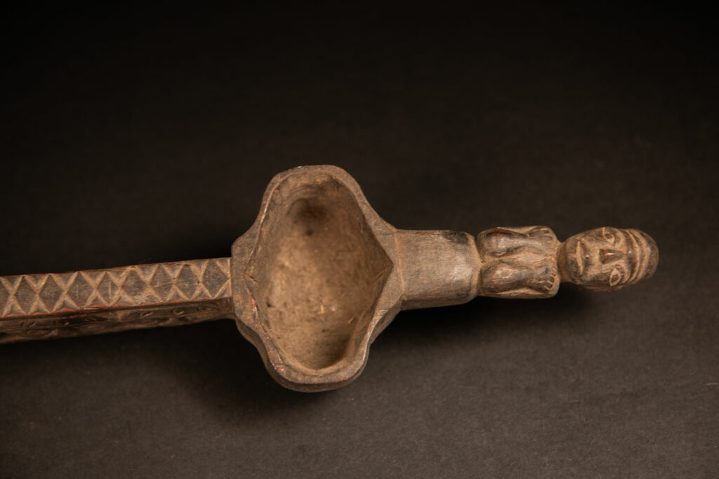 This drinking cup from paiwan features two beautifully carved handles, each depicting an ancestor holding the drinking cup between his thighs. The center of the double cup is carved with four human faces and a decorative frieze. The entire piece is made of natural wood.