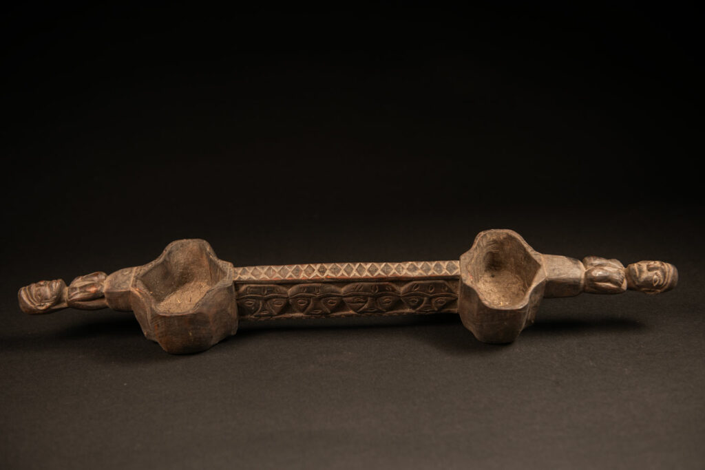 This drinking cup from paiwan features two beautifully carved handles, each depicting an ancestor holding the drinking cup between his thighs. The center of the double cup is carved with four human faces and a decorative frieze. The entire piece is made of natural wood.