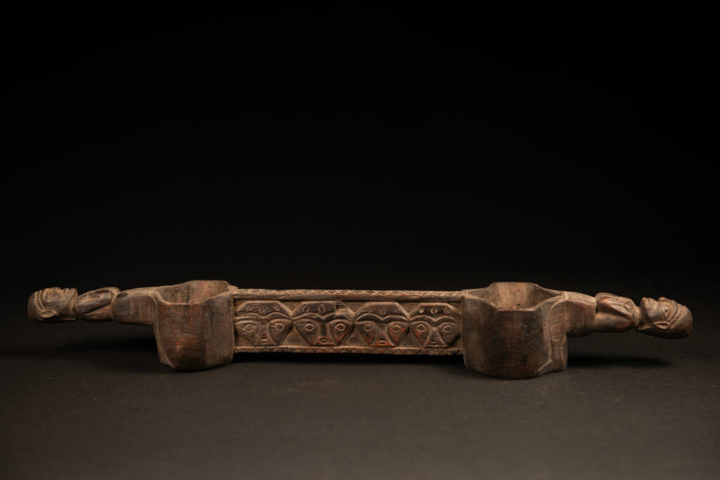 This drinking cup from paiwan features two beautifully carved handles, each depicting an ancestor holding the drinking cup between his thighs. The center of the double cup is carved with four human faces and a decorative frieze. The entire piece is made of natural wood.
