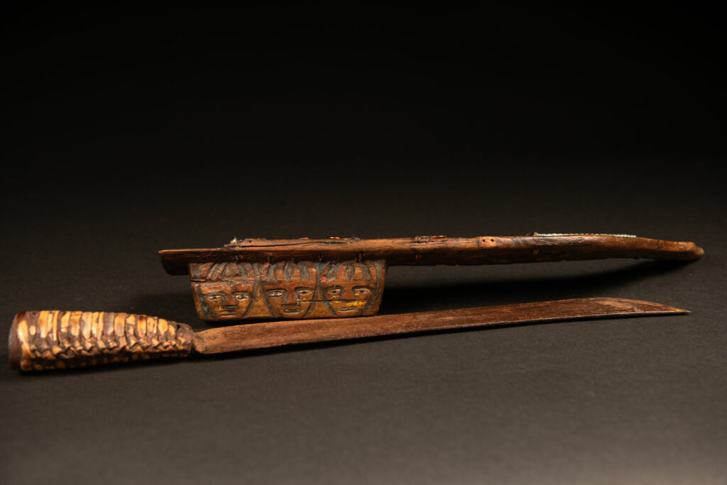 Sword from the Paiwan tribe, carved with stylized standing human figures, a serpent, and three faces. The blade and handle are forged from a single piece of iron.