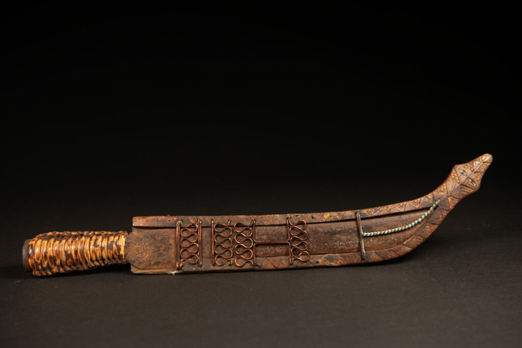 Sword from the Paiwan tribe, carved with stylized standing human figures, a serpent, and three faces. The blade and handle are forged from a single piece of iron.