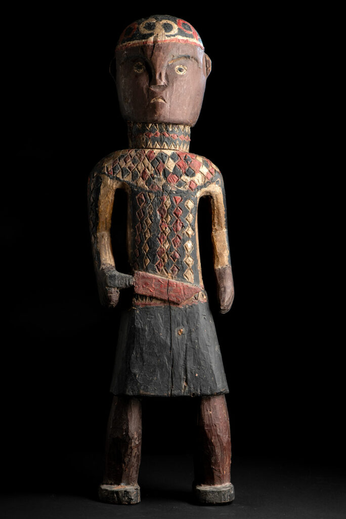 Wooden Statue from the Paiwan tribe painted in red, beige, and black, depicting a full-standing ancestor wearing ceremonial clothing and a leather headdress, with a knife at the waist.