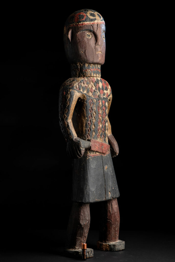 Wooden Statue from the Paiwan tribe painted in red, beige, and black, depicting a full-standing ancestor wearing ceremonial clothing and a leather headdress, with a knife at the waist.