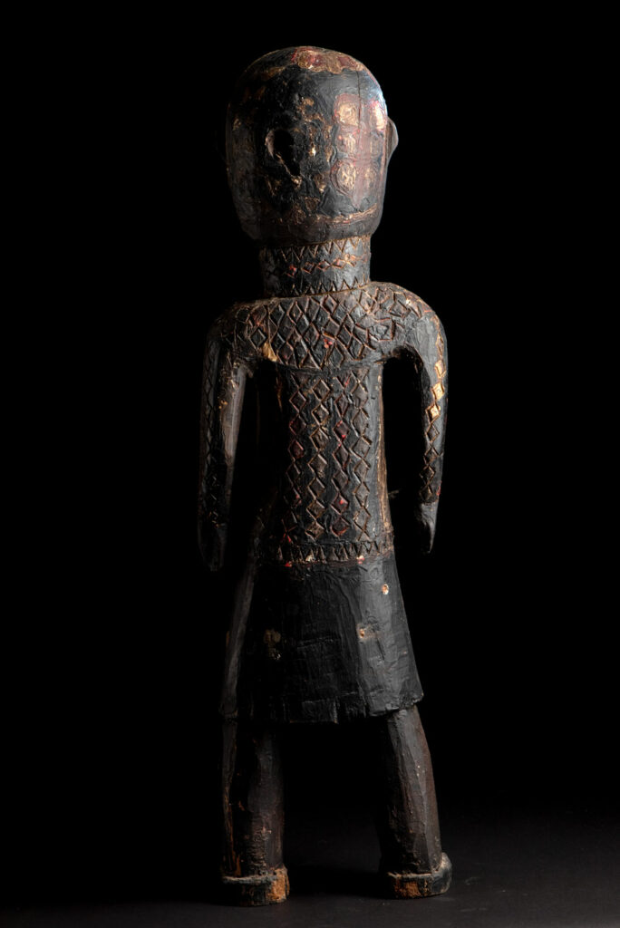Wooden Statue from the Paiwan tribe painted in red, beige, and black, depicting a full-standing ancestor wearing ceremonial clothing and a leather headdress, with a knife at the waist.