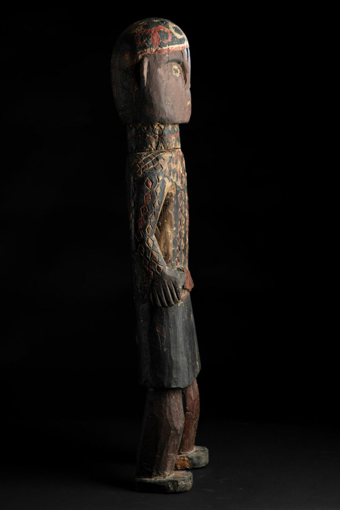 Wooden Statue from the Paiwan tribe painted in red, beige, and black, depicting a full-standing ancestor wearing ceremonial clothing and a leather headdress, with a knife at the waist.