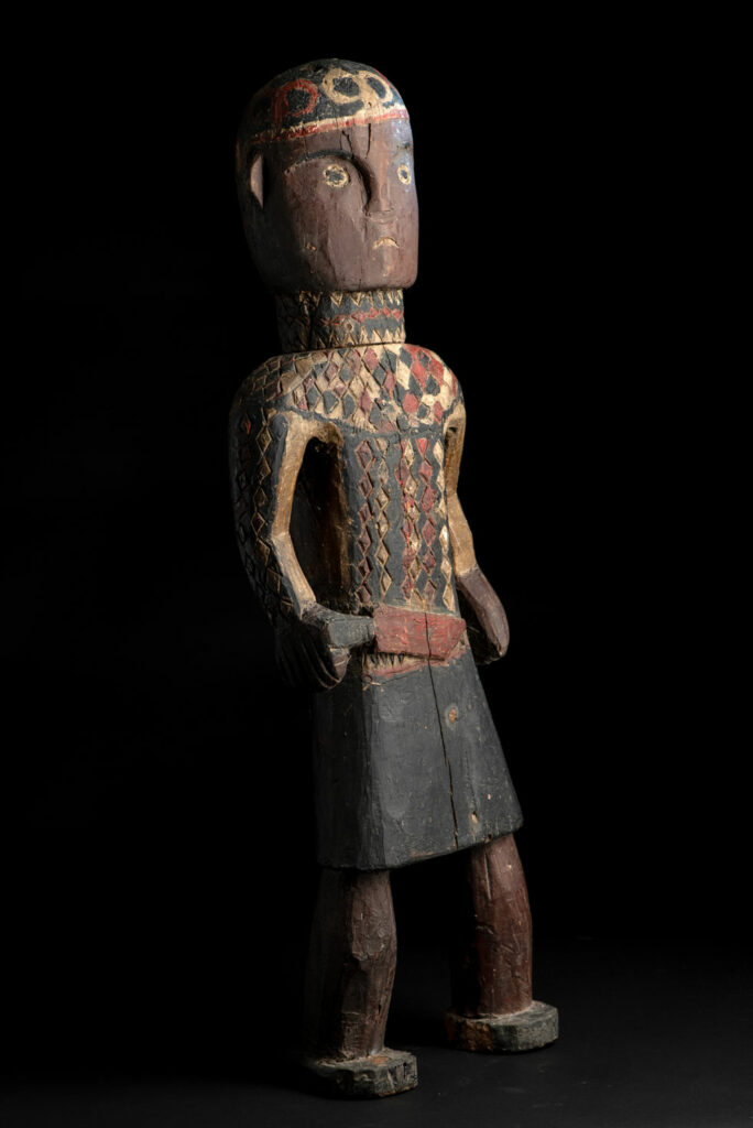Wooden Statue from the Paiwan tribe painted in red, beige, and black, depicting a full-standing ancestor wearing ceremonial clothing and a leather headdress, with a knife at the waist.