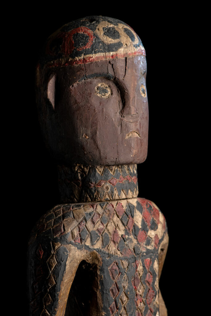 Wooden Statue from the Paiwan tribe painted in red, beige, and black, depicting a full-standing ancestor wearing ceremonial clothing and a leather headdress, with a knife at the waist.