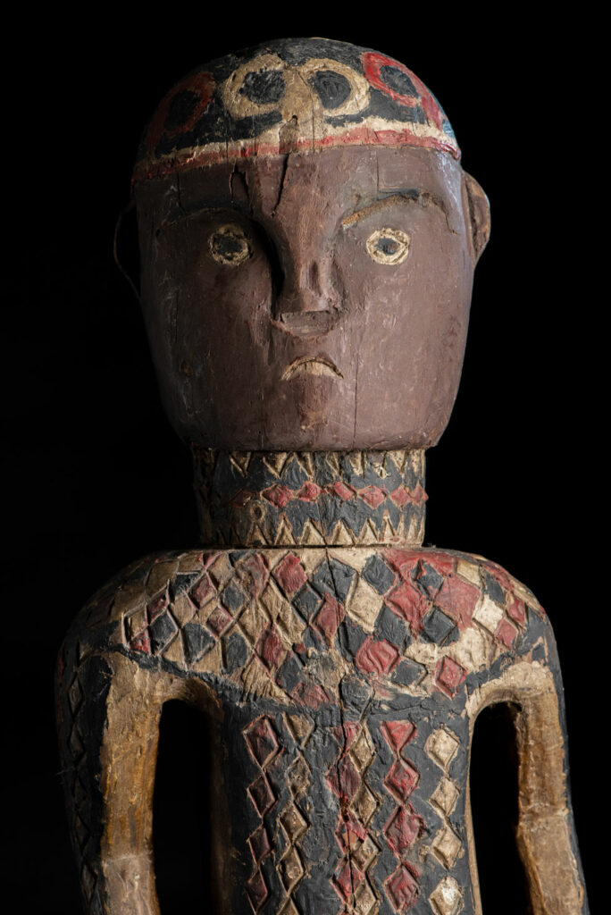 Wooden Statue from the Paiwan tribe painted in red, beige, and black, depicting a full-standing ancestor wearing ceremonial clothing and a leather headdress, with a knife at the waist.