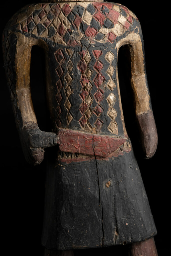 Wooden Statue from the Paiwan tribe painted in red, beige, and black, depicting a full-standing ancestor wearing ceremonial clothing and a leather headdress, with a knife at the waist.