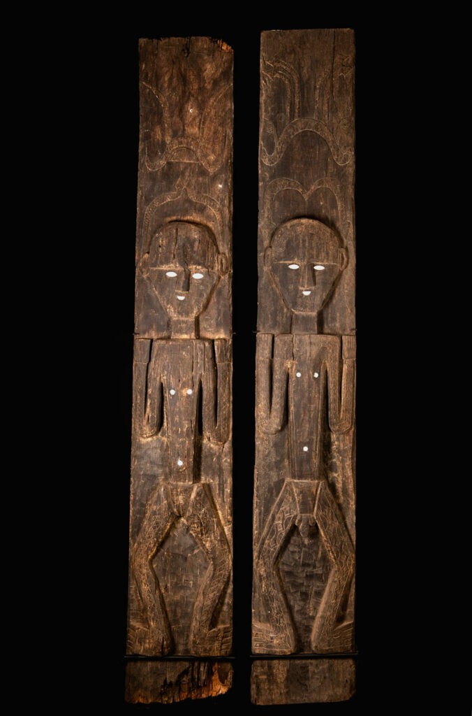 Pair of Paiwan Tribe Ancestor Panels, likely from Laiyi, carved with two figures—a male and a female. Their eyes, mouth, nipples, and navel are inlaid with porcelain. Their arms and legs are decorated with tribal motifs suggesting tattoos. Both figures wear hats, and the snakes carved above their heads indicate that they were important members of the nobility.