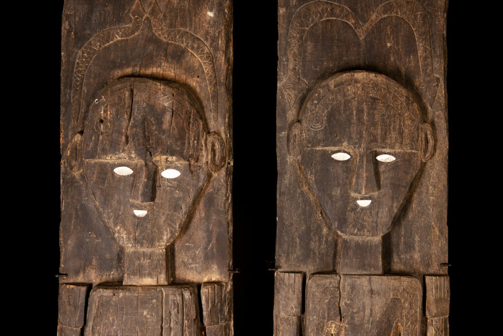 Pair of Paiwan Tribe Ancestor Panels, likely from Laiyi, carved with two figures—a male and a female. Their eyes, mouth, nipples, and navel are inlaid with porcelain. Their arms and legs are decorated with tribal motifs suggesting tattoos. Both figures wear hats, and the snakes carved above their heads indicate that they were important members of the nobility.