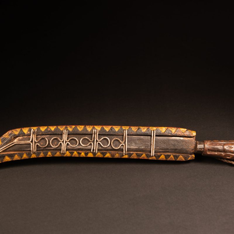 Hunting sword from the Paiwan tribe, carved with a snake and a decorative frieze on the front, as well as a stylized standing figure, a human face, and a snake. The side of the scabbard is carved with two faces. The entire piece is painted in red, brown, white, black, and yellow. The paint was likely applied at a later time.