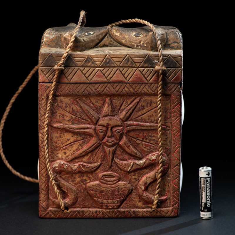 Ancient shaman box from the Paiwan tribe, stained red, with a sliding lid carved with snakes face. One side is carved with two tattooed hands, while the other features a sun, a pot, and two snakes — depicting the myth of the Paiwan people's origin from the union of a pot and a snake. The sides are inlaid with circular shell ornaments.